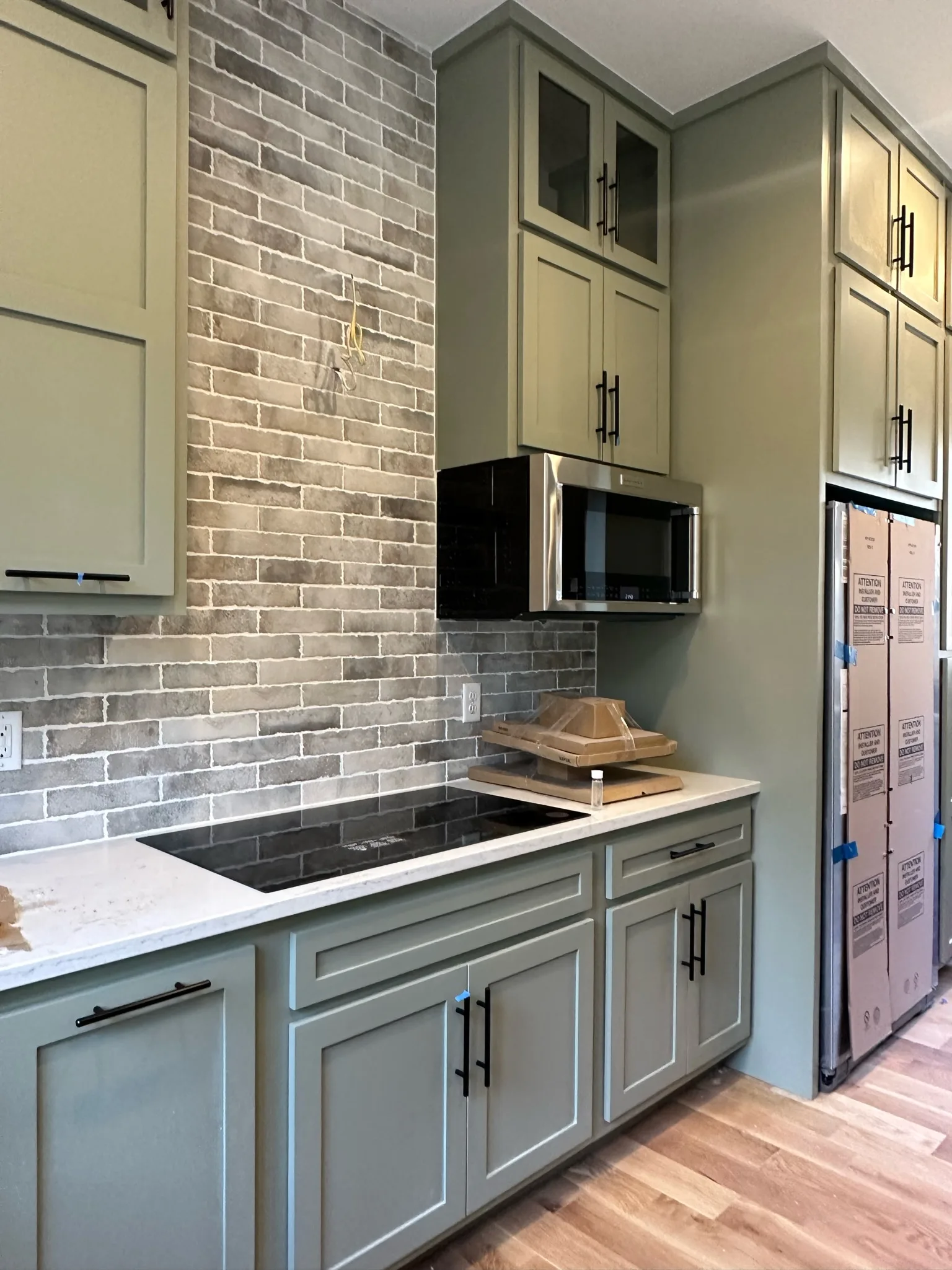 Sage green shaker kitchen cooktop wall with brick backsplash by Sims Custom Woodwork.