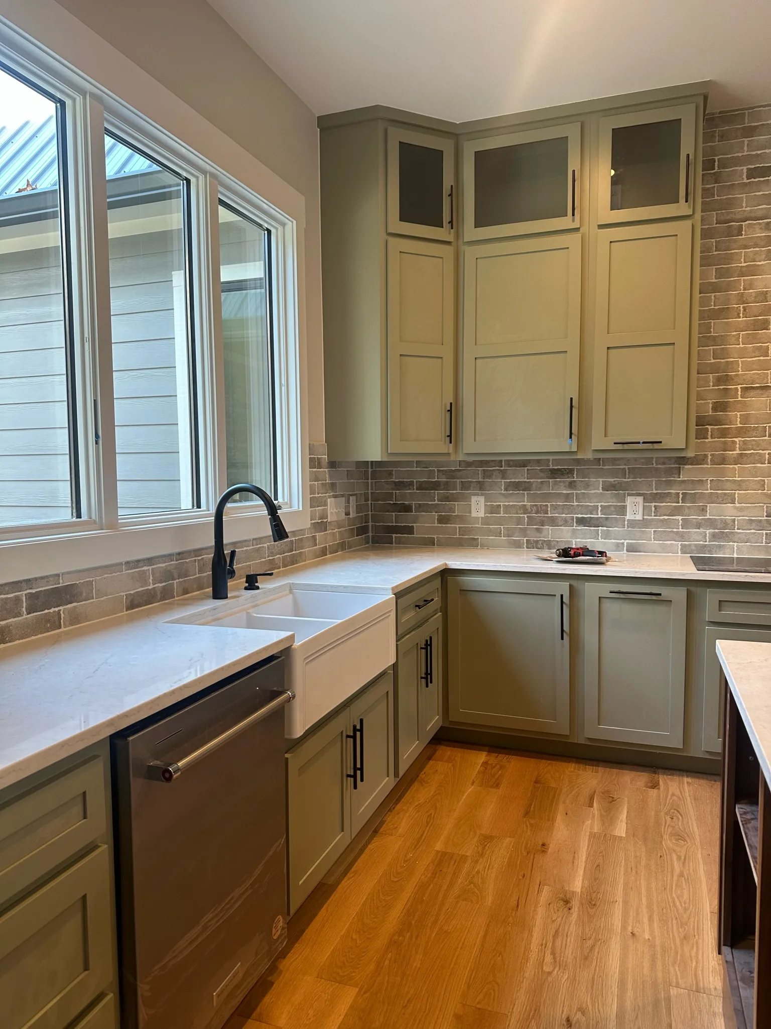 Corner sink view of sage green shaker kitchen with farmhouse sink and stone backsplash by Sims Custom Woodwork.